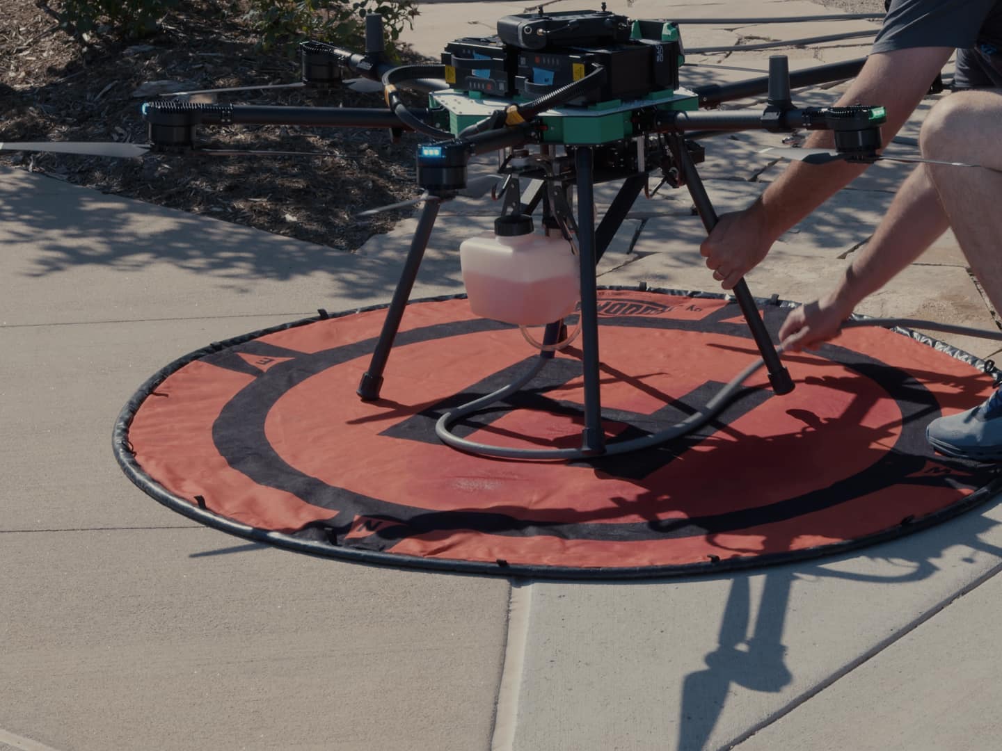 A crew member checks a cleaning drone on an orange landing pad, with hands on its leg and hose.