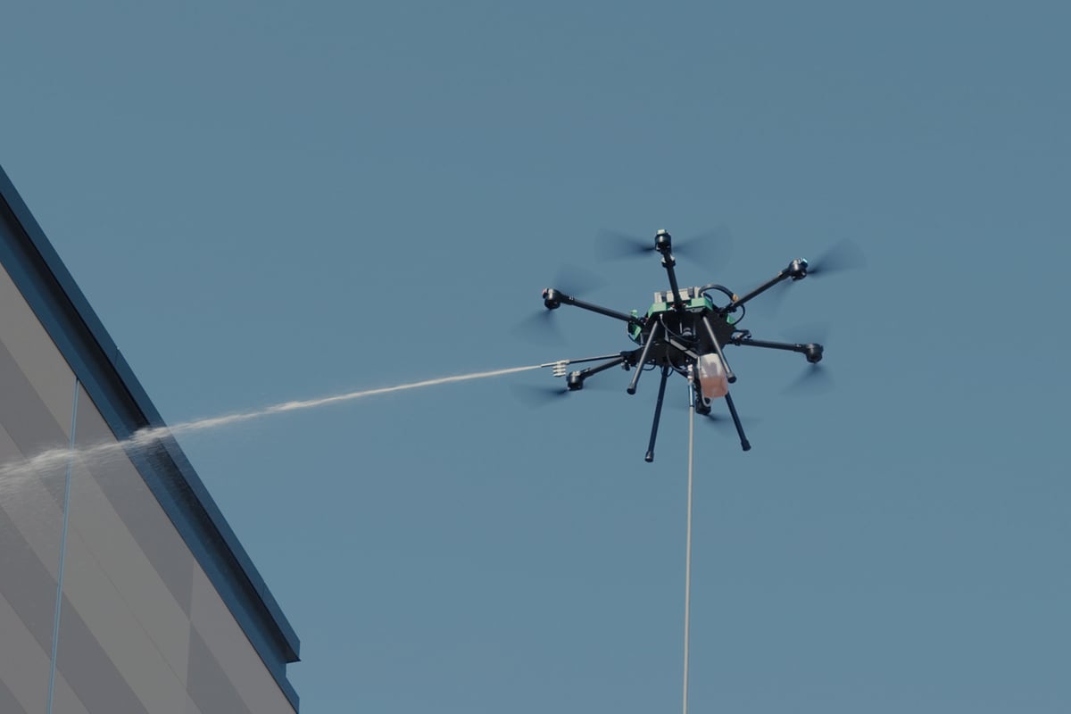 A tethered hexacopter cleaning drone hovers against a clear blue sky and sprays a long jet of water toward a building at its left; its hose hangs straight down.