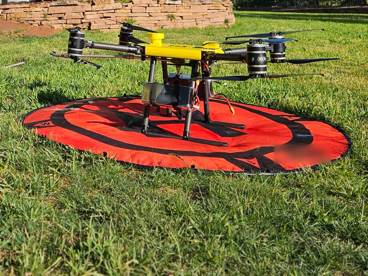 Seen from low on the lawn, a yellow-top cleaning drone rests on its orange landing pad, a stone wall behind it.