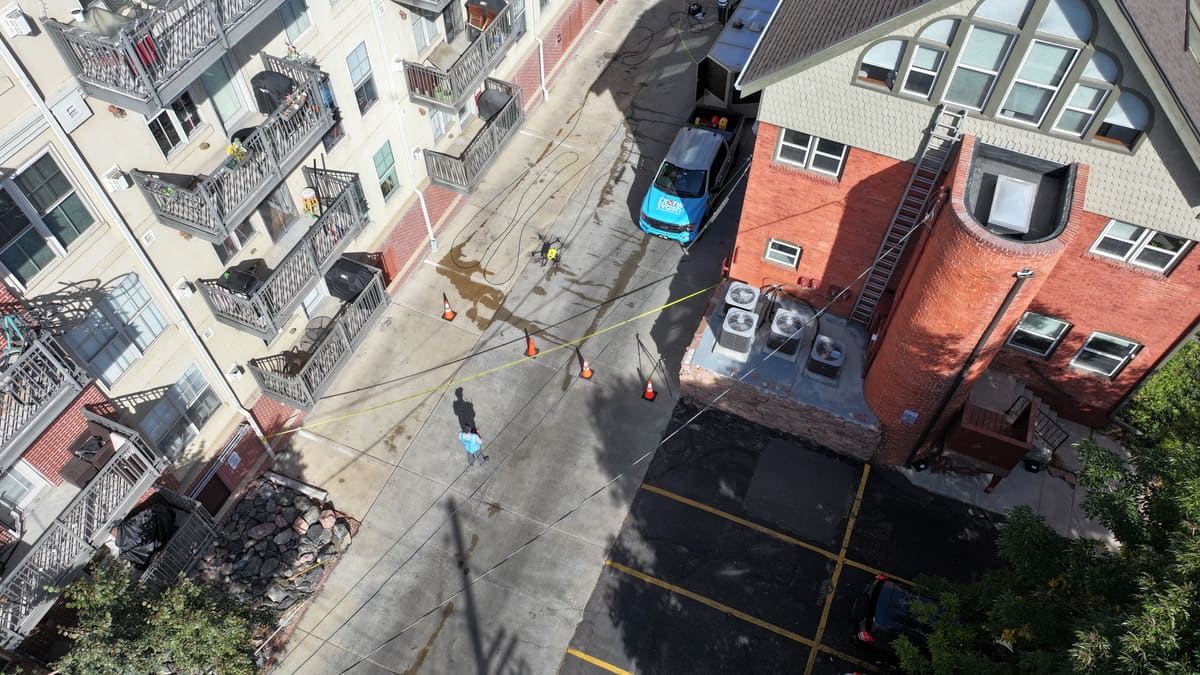 From above: a crew member in a company vest at the controls on a drive closed with caution tape and cones, our cleaning drone over the pavement and our truck beside the hose lines.