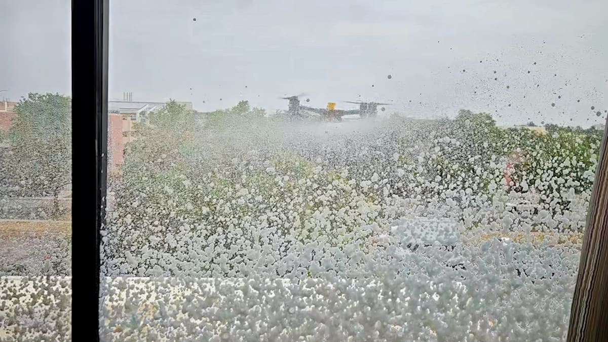 Seen from inside through a window, white cleaning foam covers the glass while a yellow-topped drone hovers beyond it above trees and low rooftops under a gray sky.