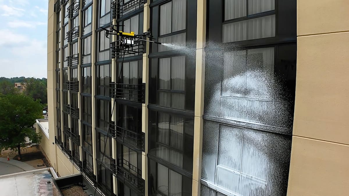 A yellow-topped cleaning drone on a thin hose fans white foam across the dark-framed windows and iron balconettes of a mid-rise building with tan pilasters, trees and sky at its left.