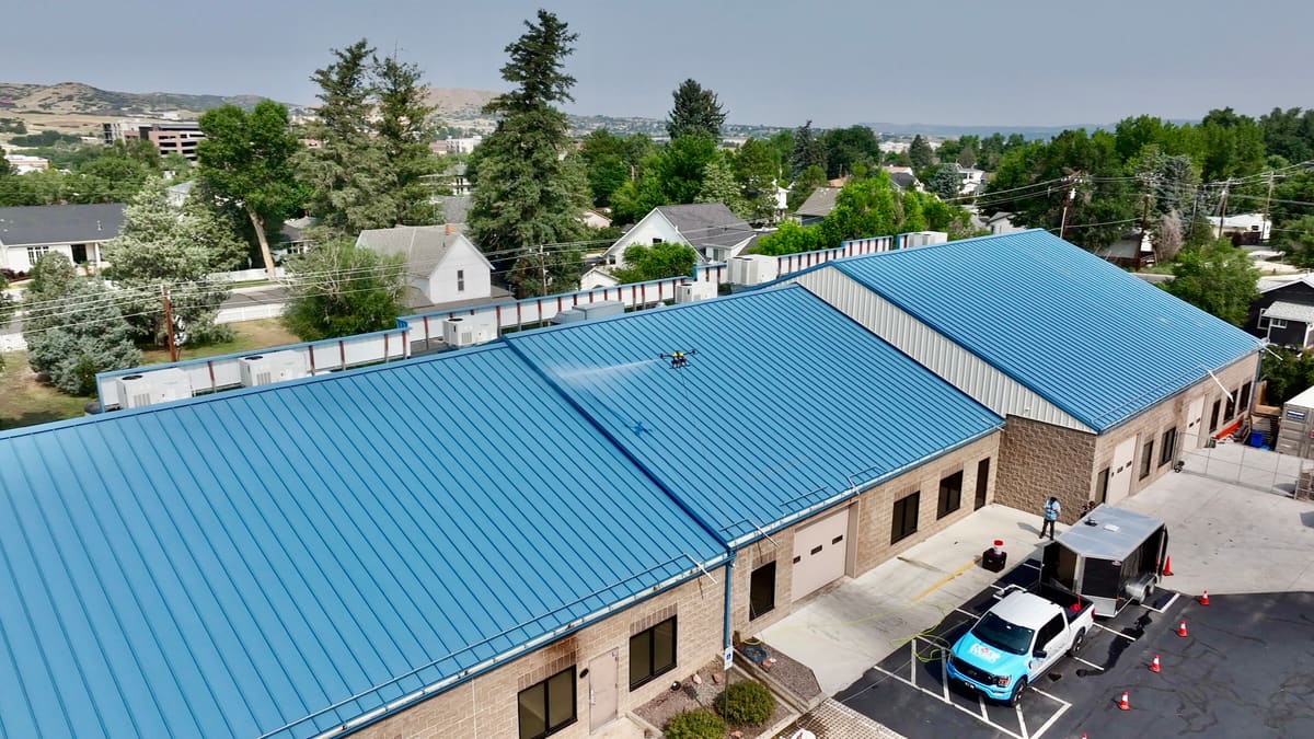 From above, one of our cleaning drones sprays a blue standing-seam metal roof on a long stone-block building, with our branded pickup, a trailer, orange cones and a person in a safety vest in the lot below.