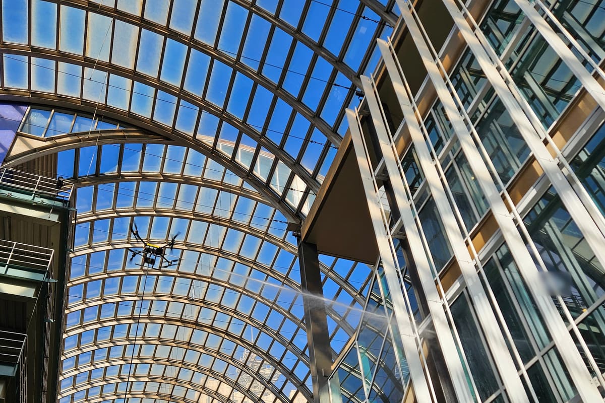 Looking up inside a glass-roofed galleria: one of our cleaning drones hovers under the arched glass vault and sprays a long jet at the glass and white fins of the building beside it.