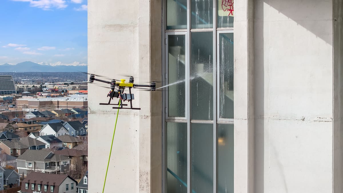 One of our yellow-topped cleaning drones hovers beside a tall concrete building and sprays a jet of water across its window panes, a lime-green hose running down from it; rooftops and snow-capped mountains stretch out behind.