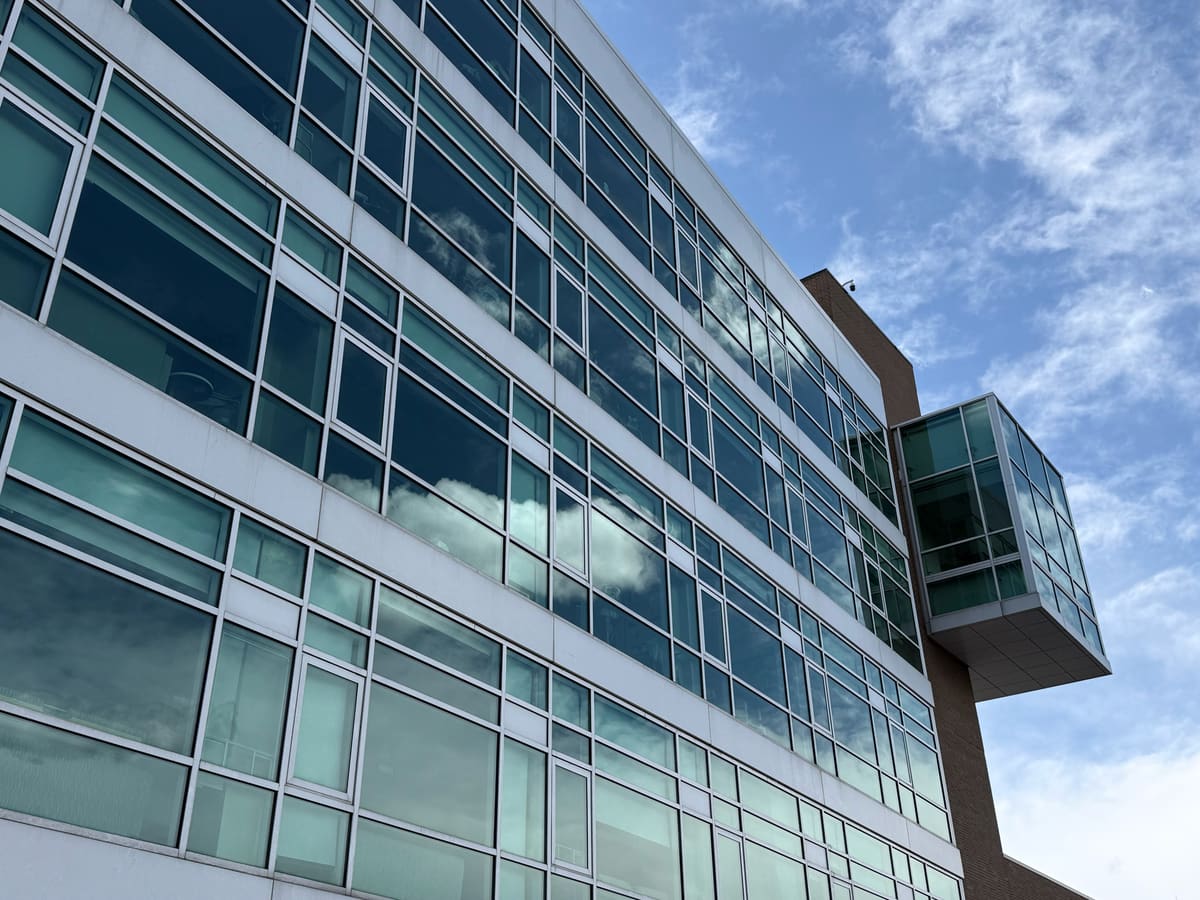 Looking up at long bands of tinted windows reflecting white clouds, a glass-walled bay jutting from a brick tower at the far end of the building, under a blue sky.