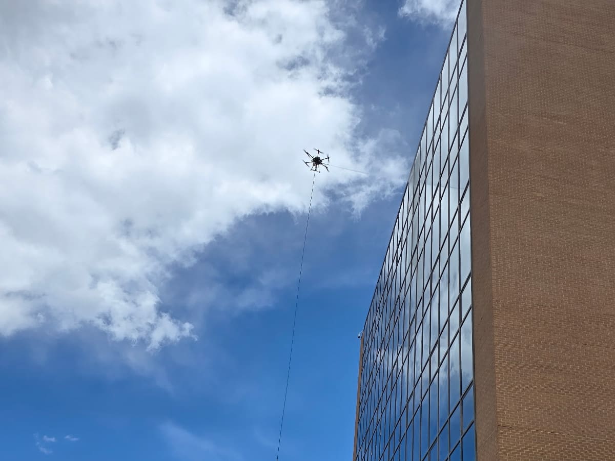 Looking up past a brick and glass tower: one of our cleaning drones hovers high against a cloudy blue sky, its hose dropping in a straight line toward the ground.
