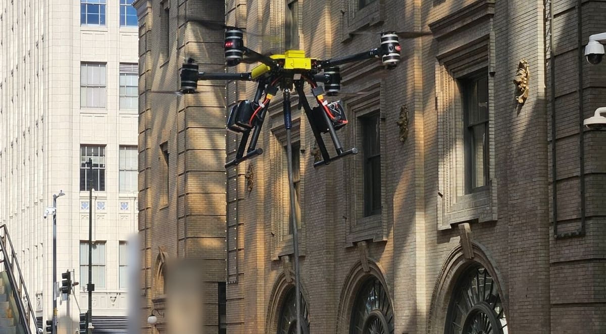 A yellow-top cleaning drone hovers on its hose beside an old stone facade with tall arched windows, a pale high-rise behind it.
