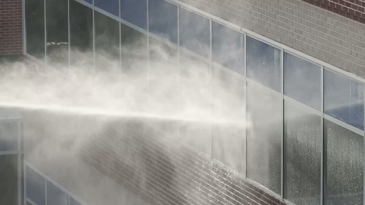 A jet of water inside a billowing white cloud of spray and steam hits the windows of a brick facade, and water runs down the glass.