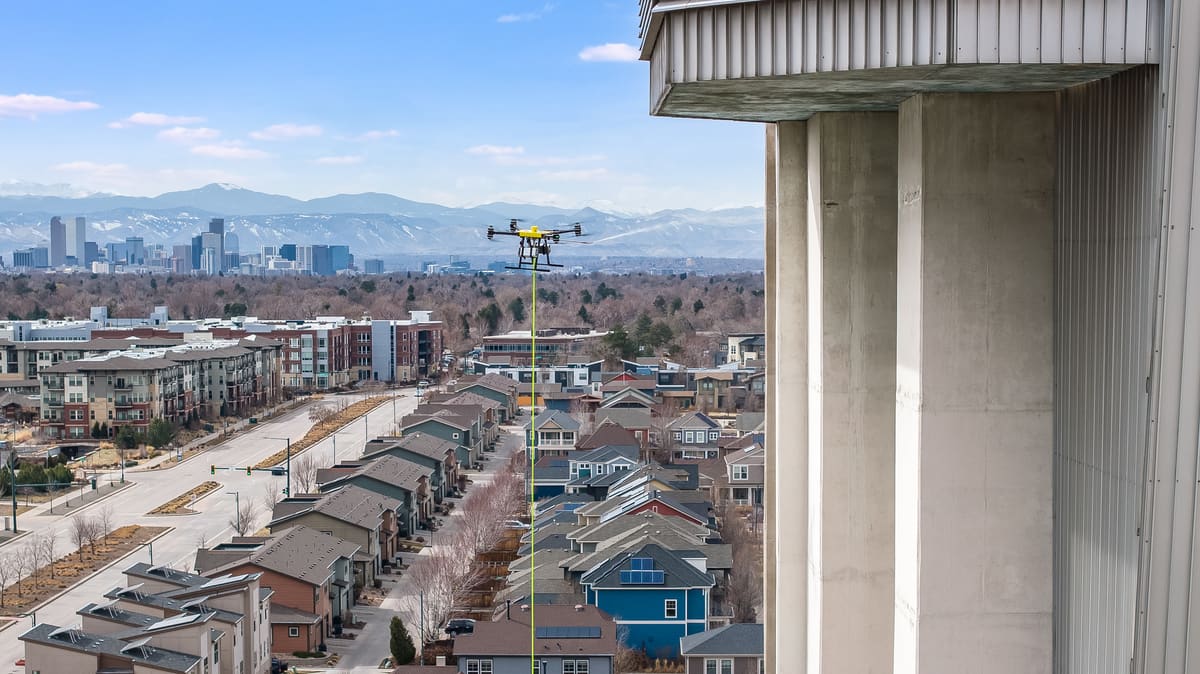 One of our cleaning drones hovers on its lime-green hose beside a weathered concrete tower, spraying toward it, with Denver's skyline and the snow-capped Front Range behind.
