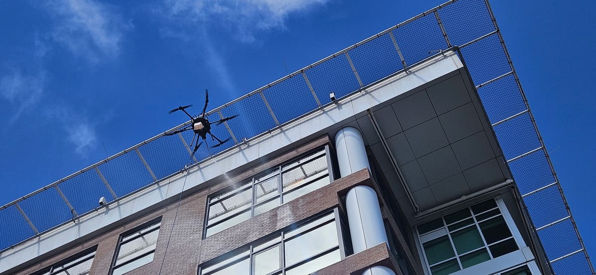 Our six-arm cleaning drone sprays the top-floor windows of a brick and glass building, just below a steel safety net that juts out from the roofline.