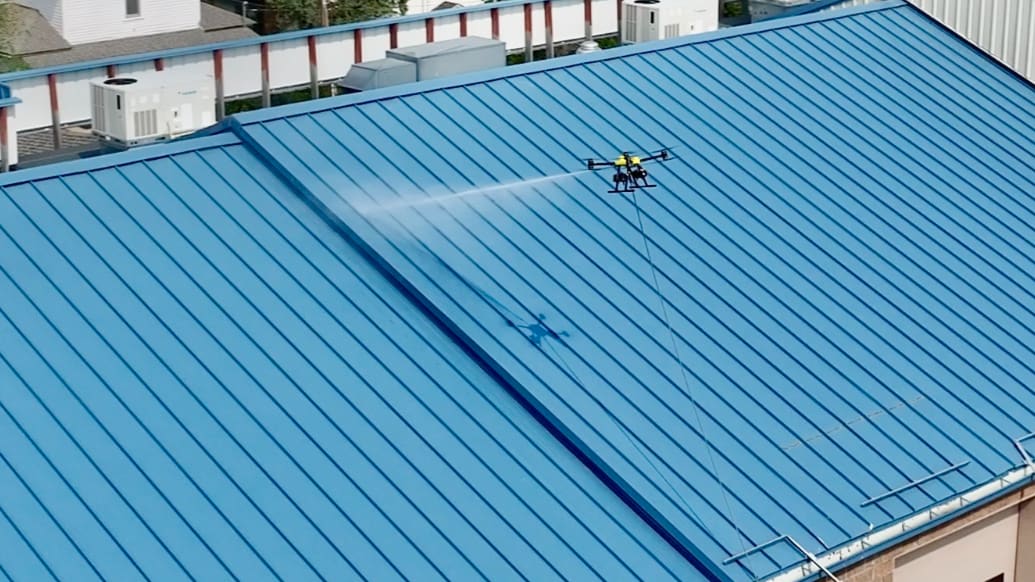 One of our cleaning drones sprays a blue standing-seam metal roof, the water fanning across the panels and its hose running down past the edge of the roof.