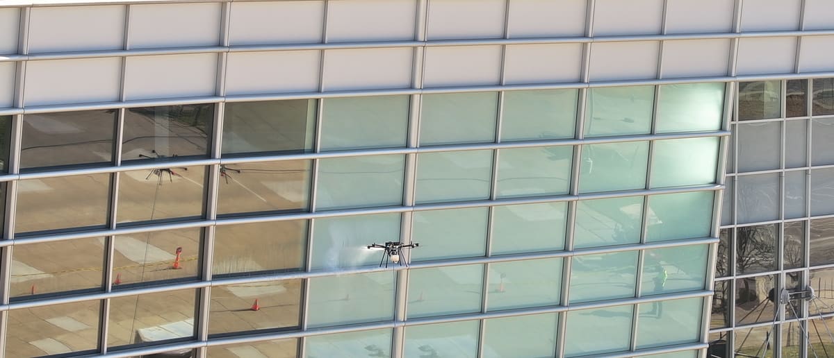 From the air, one of our tethered cleaning drones sprays a stadium's curved glass curtain wall beneath its band of white metal panels; the glass reflects the plaza, the cones and the drone itself.