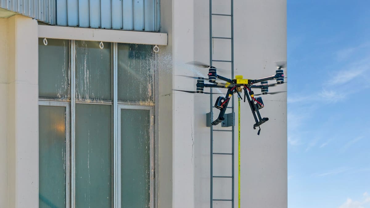 A yellow-topped cleaning drone, its hose hanging below it, sprays the tall windows of a white building beside a wall ladder, with blue sky to its right.