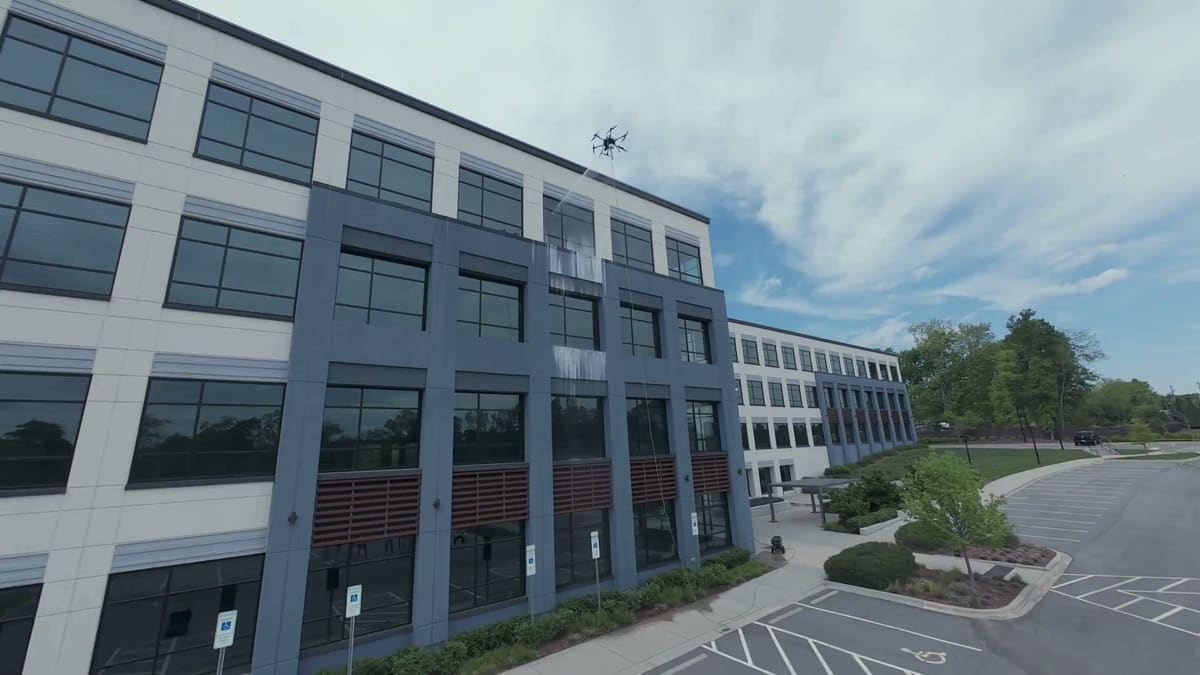 A tethered cleaning drone sprays the top-floor windows of a four-story building clad in gray and white panels, water streaking down the glass, with a parking lot and trees beside it.