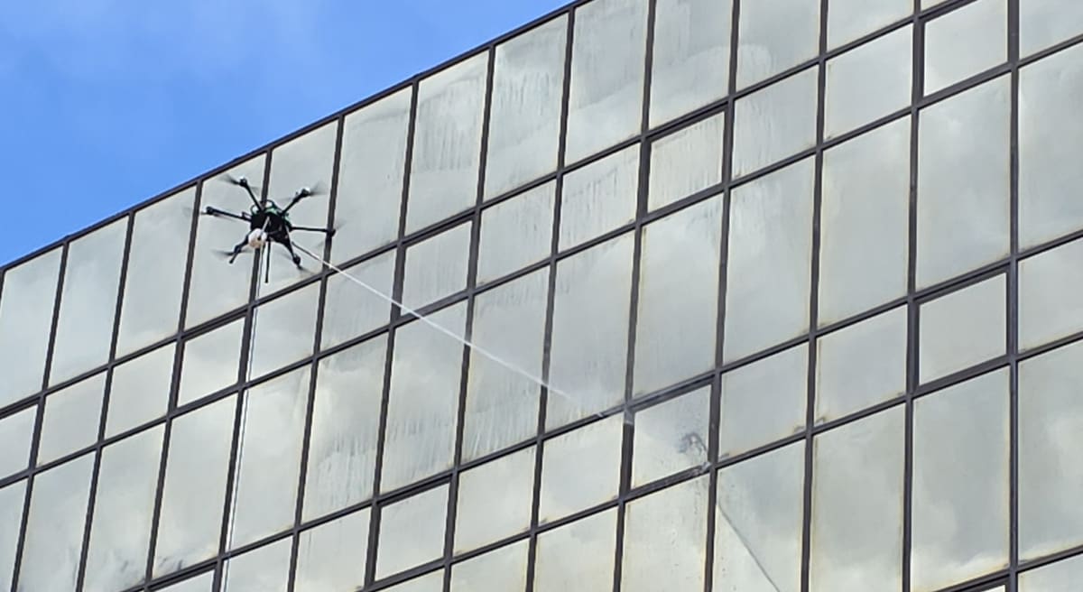One of our cleaning drones fires a long jet across a gray glass curtain wall below the roofline, the upper panes hazed and spotted, a blue sky above.