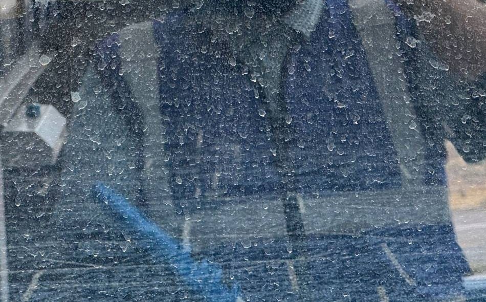 Close-up of a commercial window crusted with white mineral spots, a blurred reflection of a person in a blue vest behind the spotting.