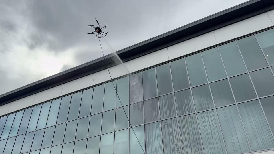 A tethered drone sprays a glass facade beneath a cloudy sky, with water running down the windows.
