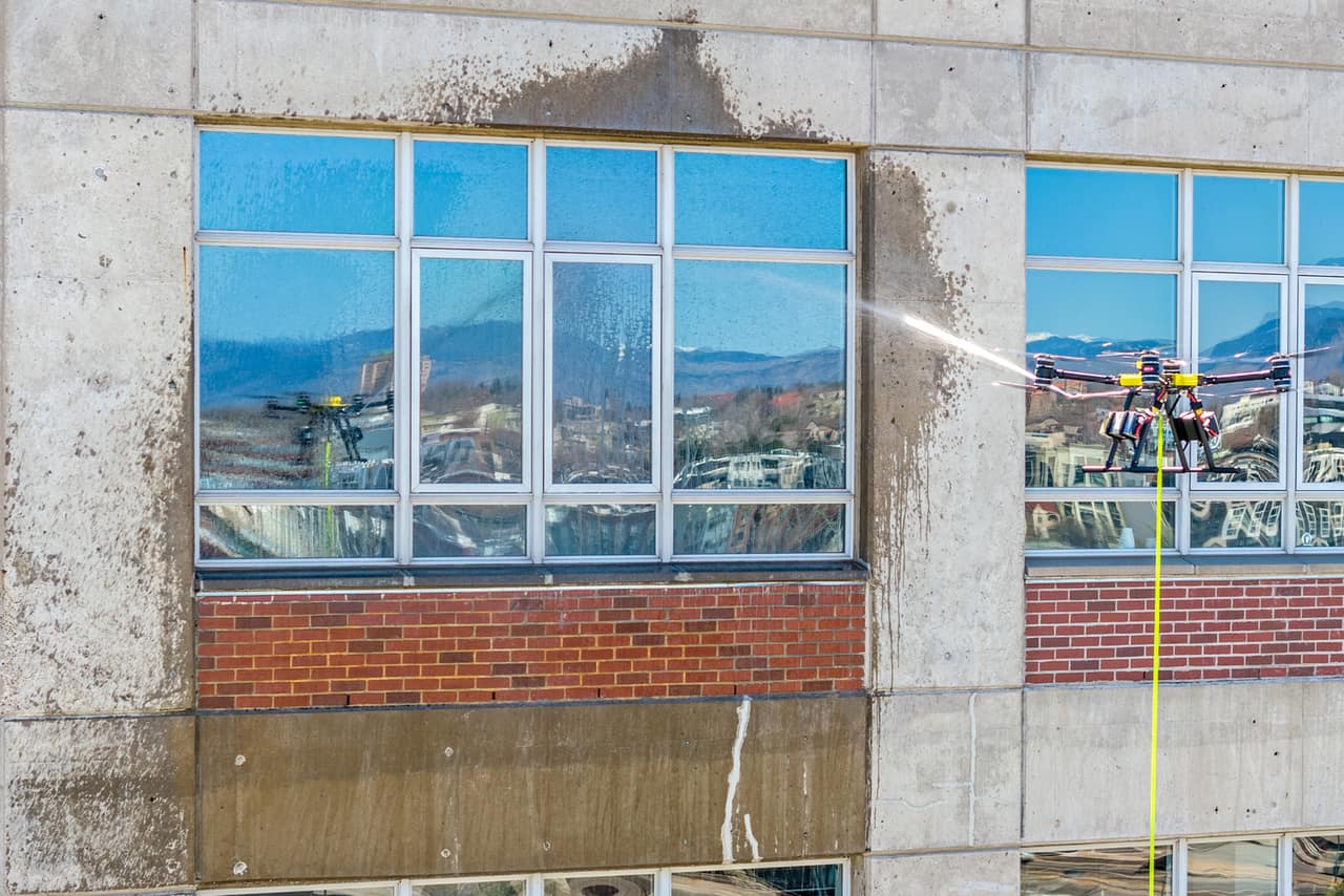 A yellow cleaning drone sprays a concrete pier beside windows reflecting mountains, with wet streaks across the facade.