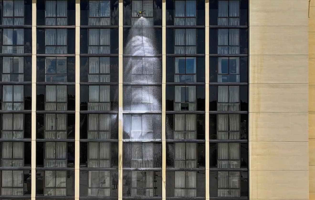 A yellow drone sprays a broad sheet of white foam down a vertical band of exterior windows.